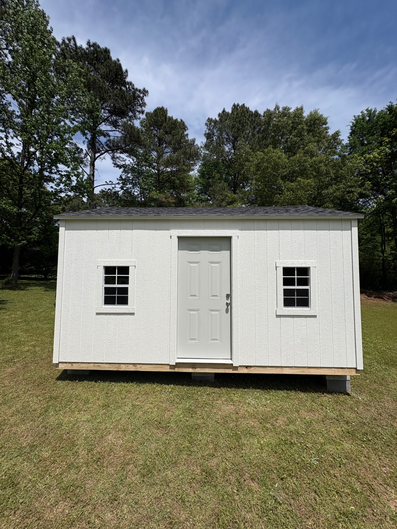 White shed with a house door between two grid windows, low shingle roof, set on blocks in a mowed yard