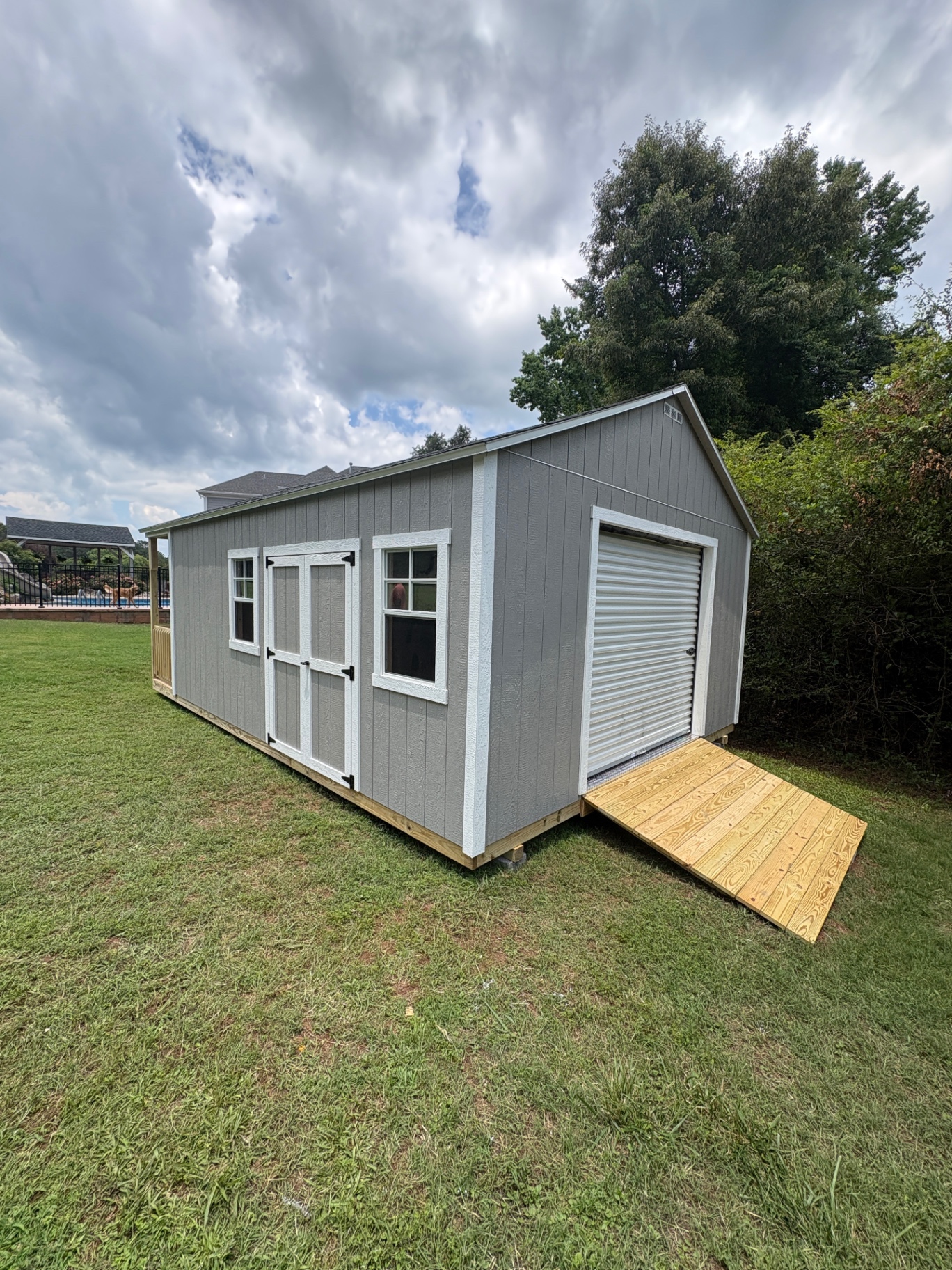 Gray shed with a white roll-up door on the gable end, a treated wood ramp, double doors and two windows along the side