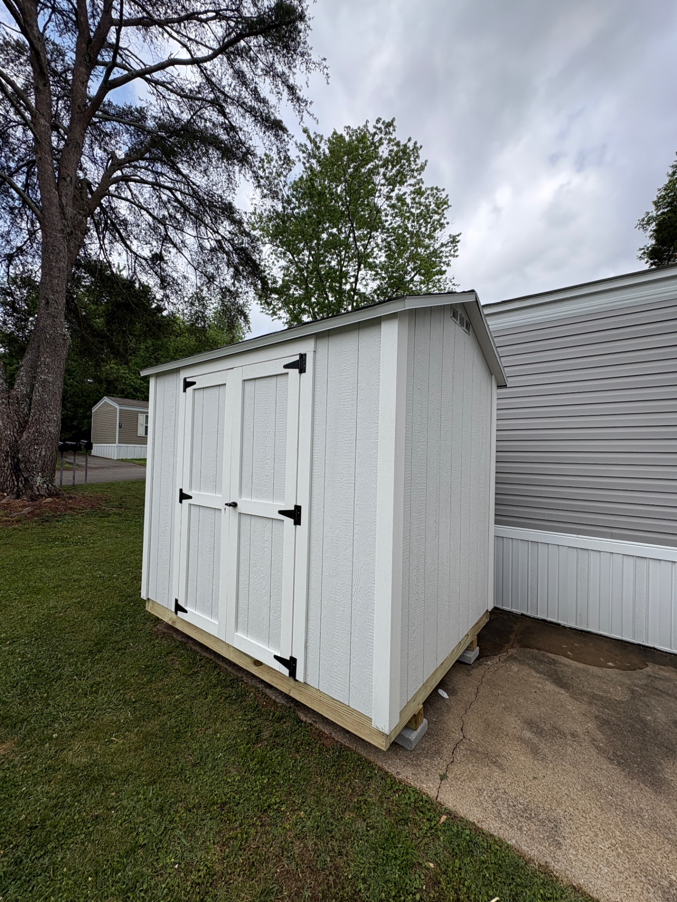 White shed with white double doors and black hinges set close beside a home on concrete blocks