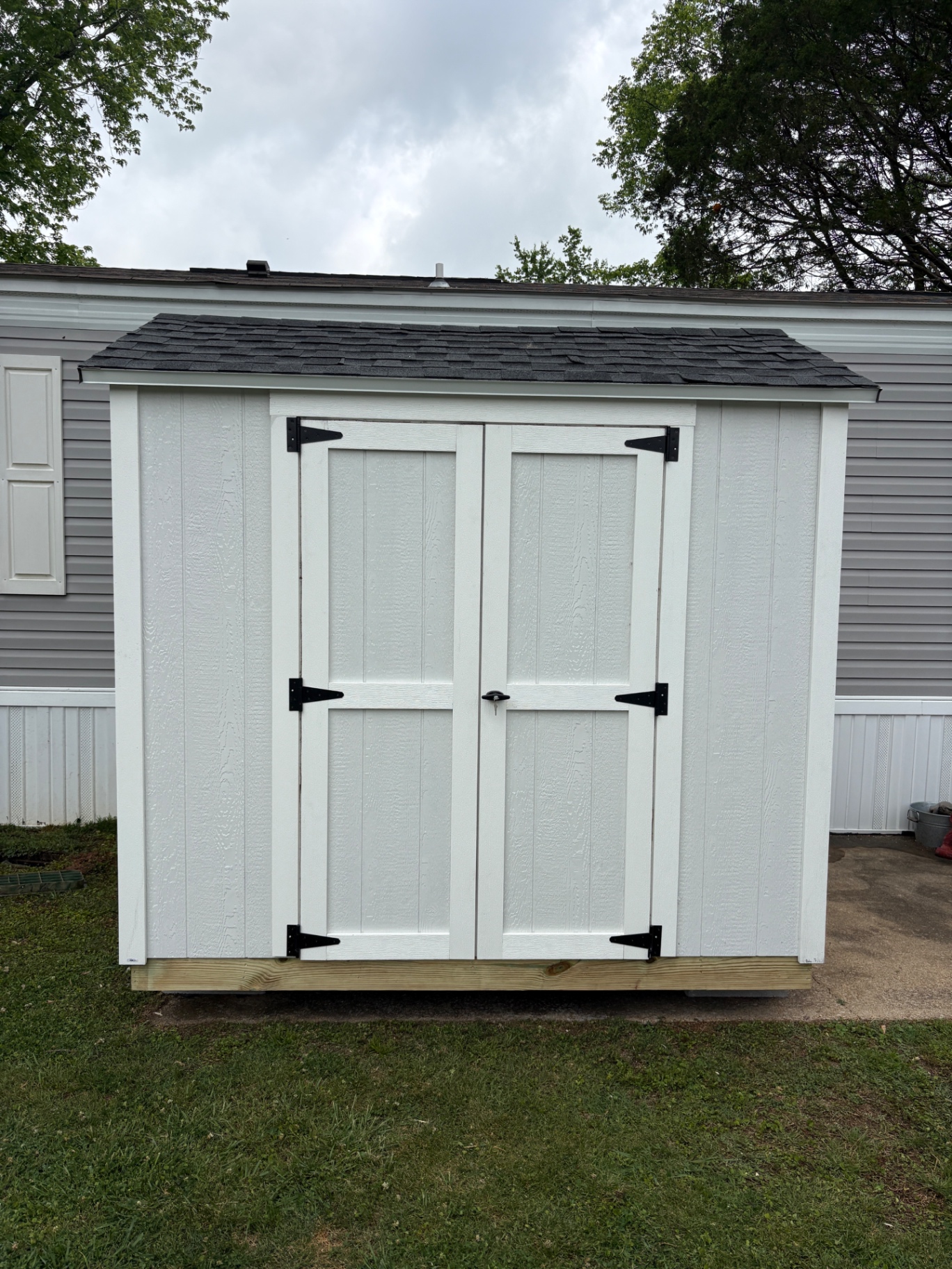 Small white shed with double doors and black hinges, tucked against a home on a concrete pad