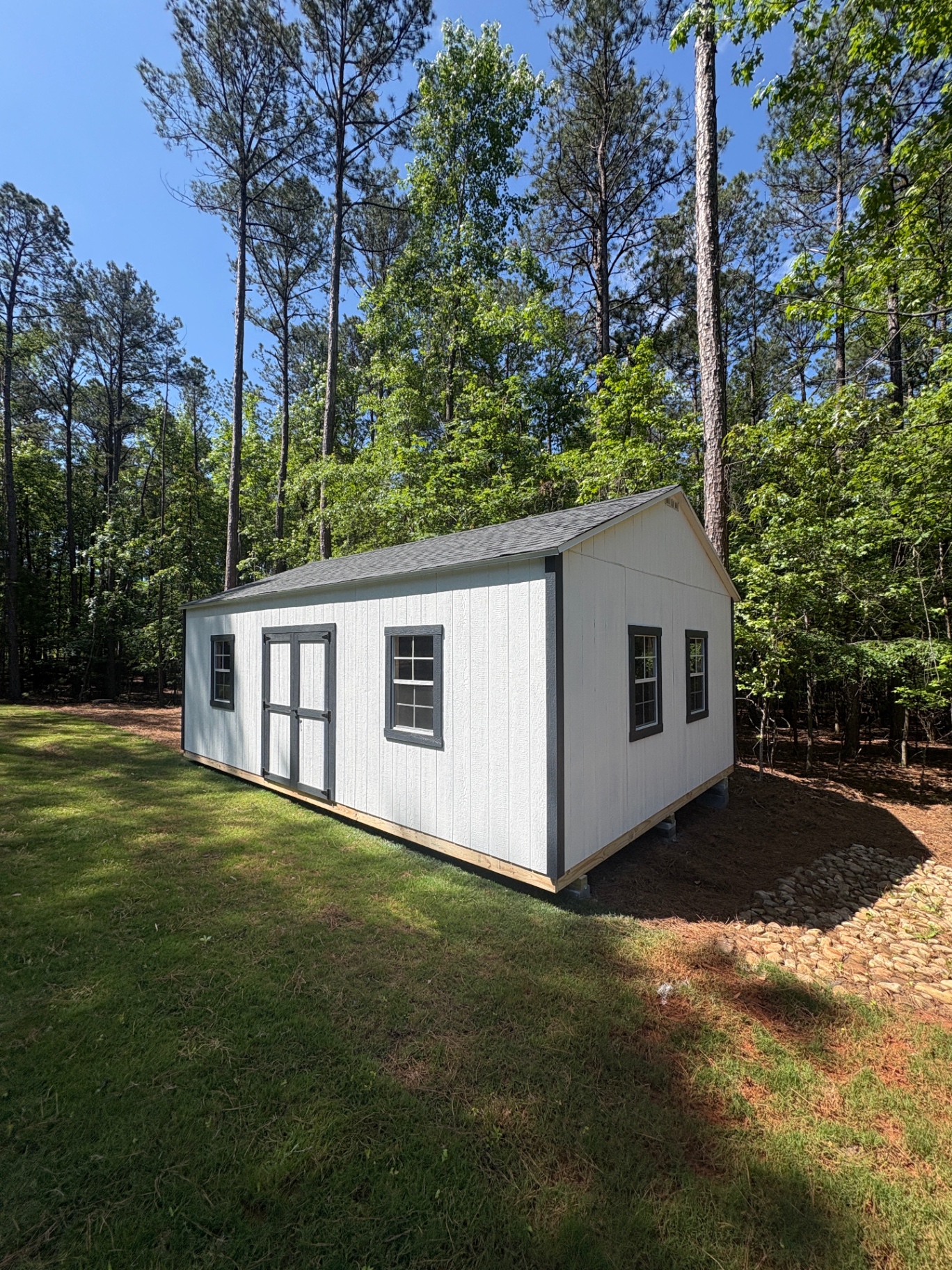 Long white shed with charcoal trim and charcoal-trimmed windows, double doors on the side and a dark shingle roof, set in pines