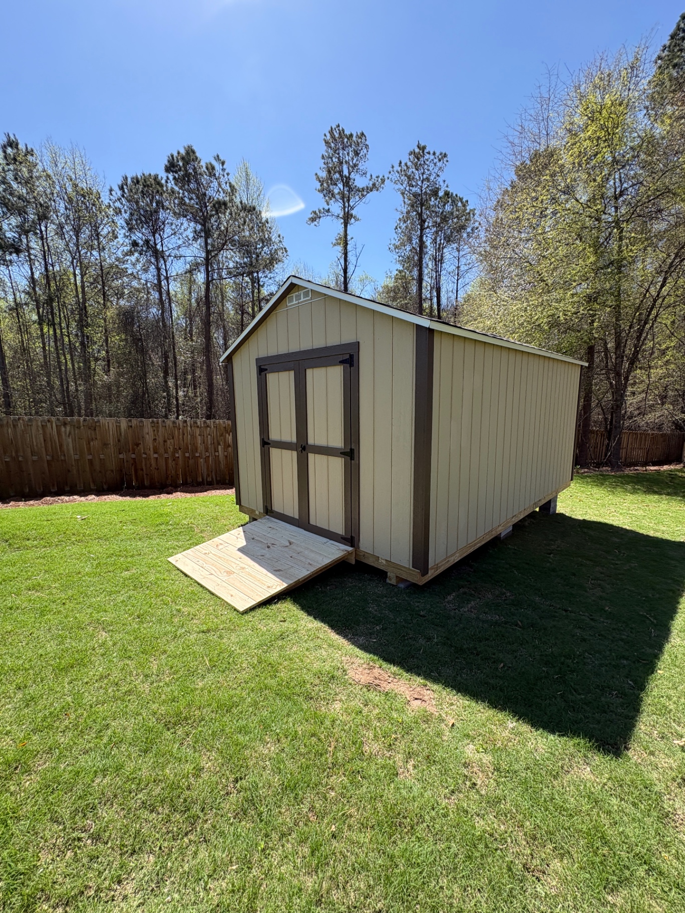 Angled view of a tan shed with dark-trimmed double doors and a treated wood ramp on a green lawn