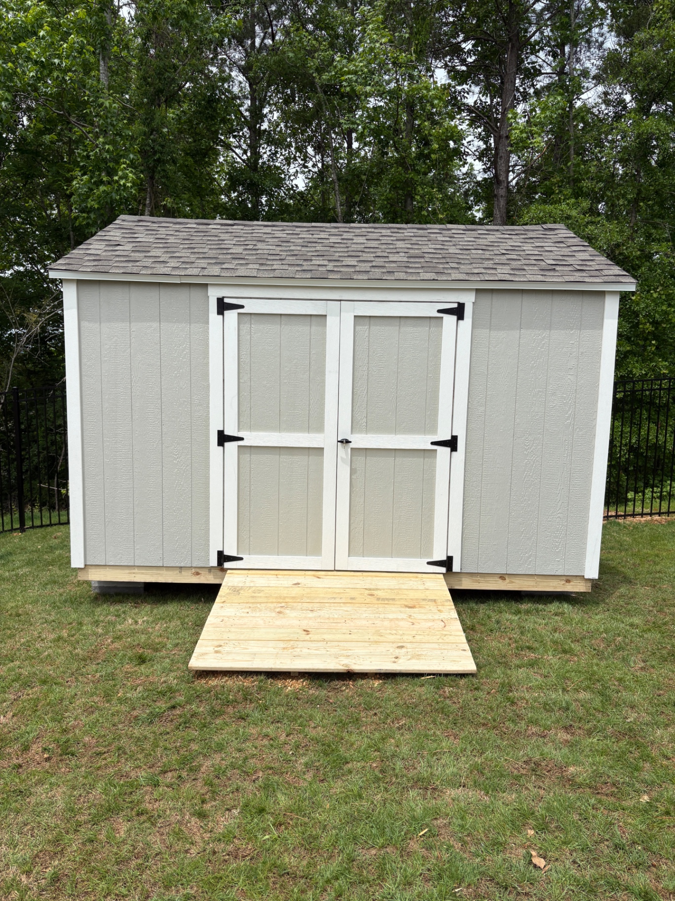 Light gray shed with white-trimmed double doors, brown shingle roof, and a wide wood ramp in a fenced yard