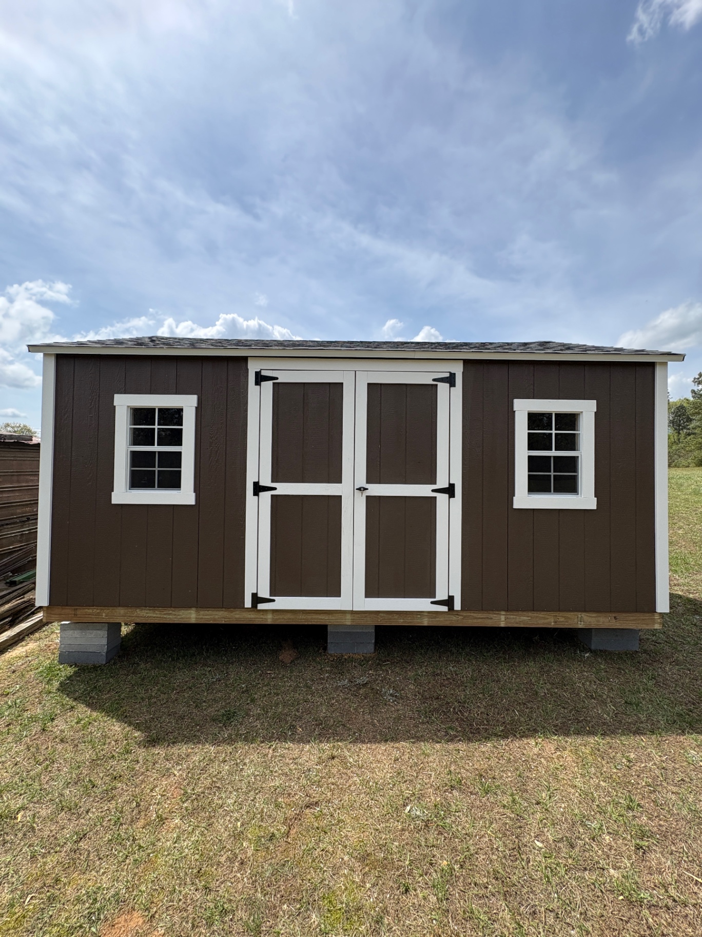 Brown shed with white trim, double doors in the center and a grid window on each side, set on concrete blocks