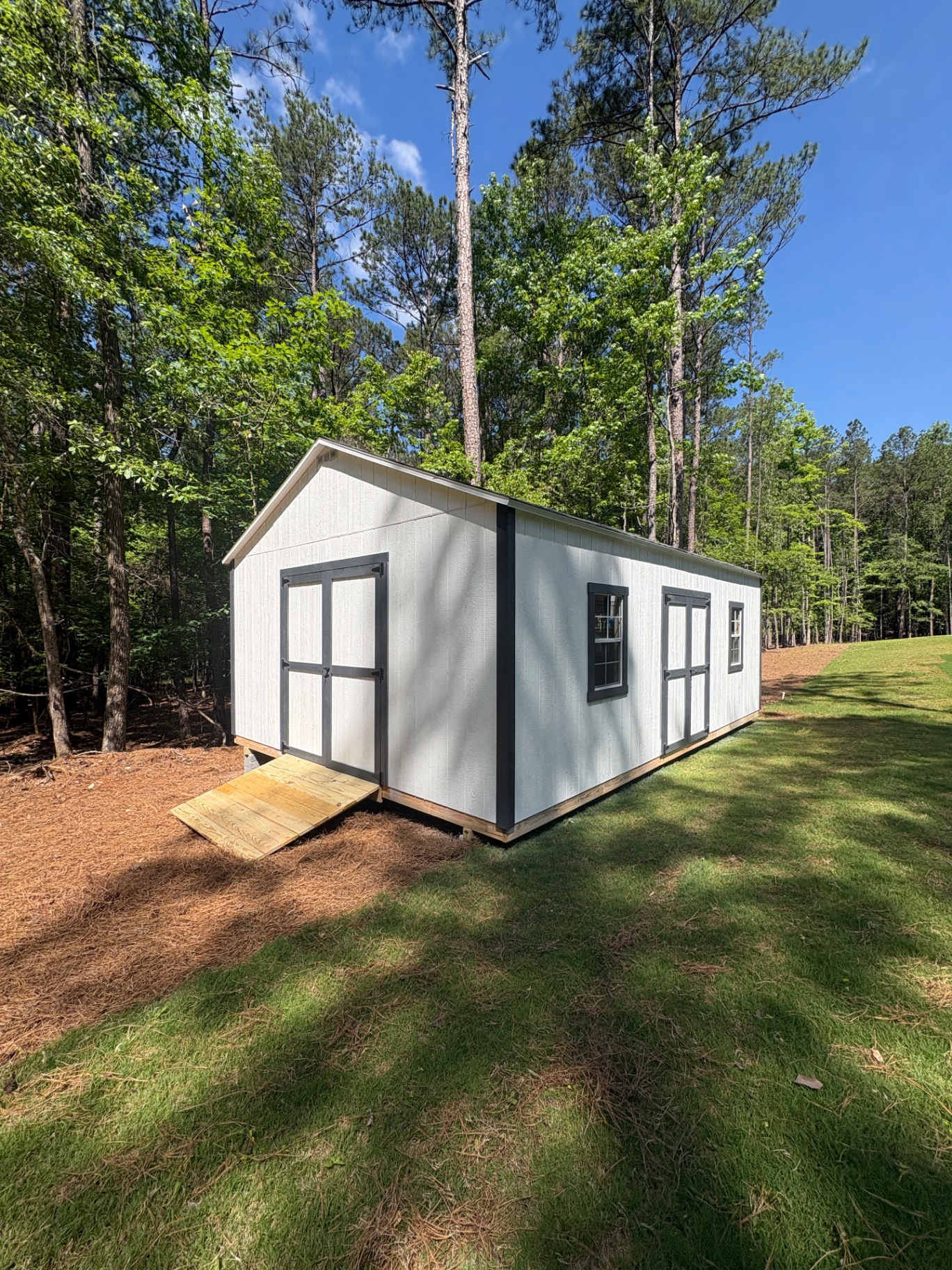 White shed with charcoal-trimmed double doors on the gable end, a second set of doors and windows on the side, and a wood ramp