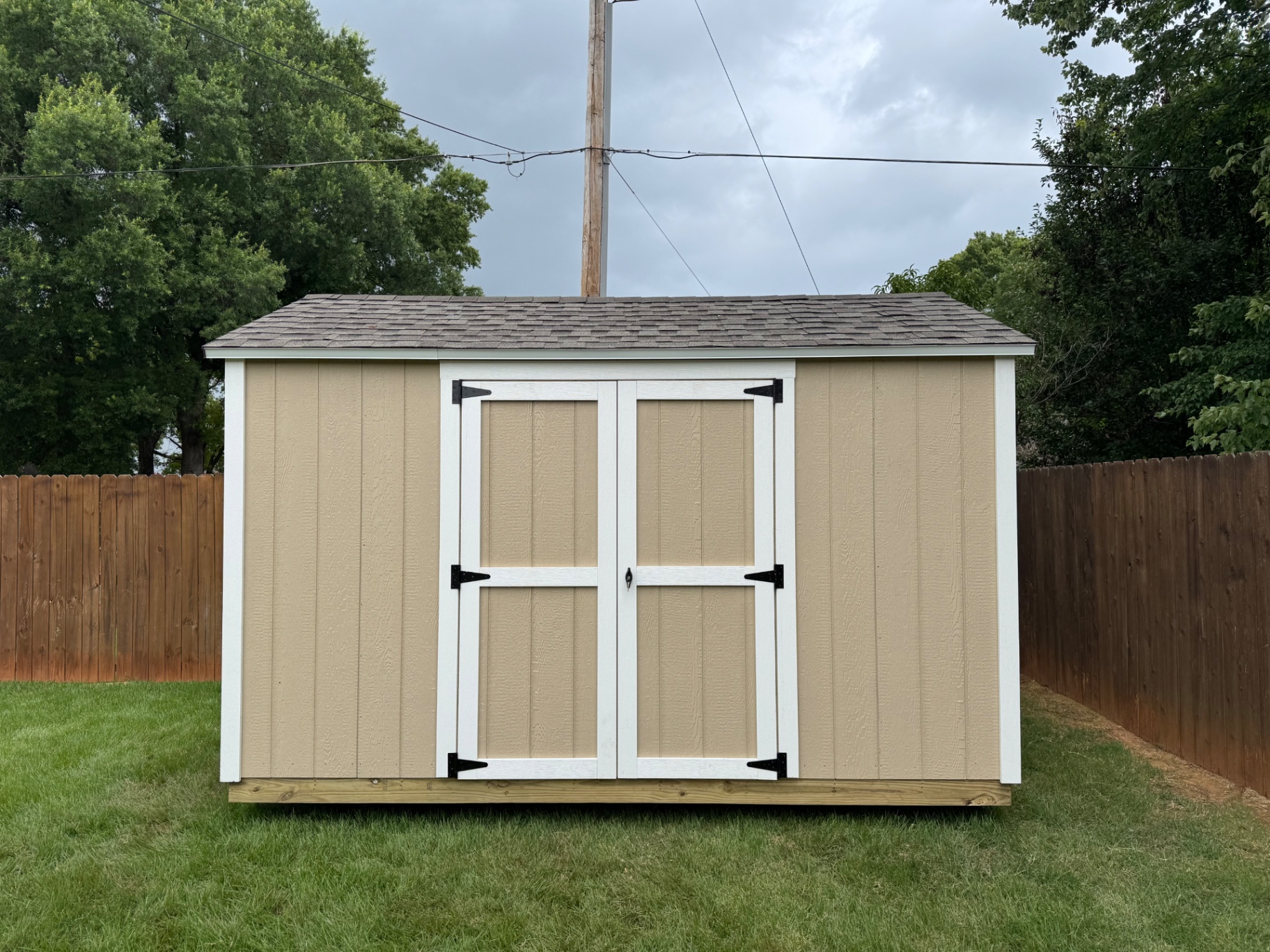 Tan shed with white-trimmed double doors and black strap hinges in a fenced backyard