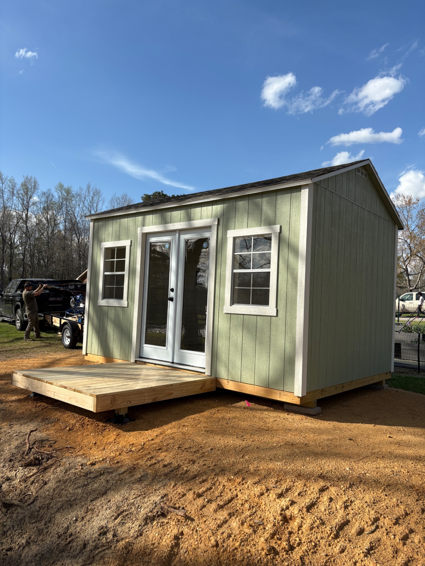 Angled view of a sage green shed with white French doors, two grid windows and a low wood platform