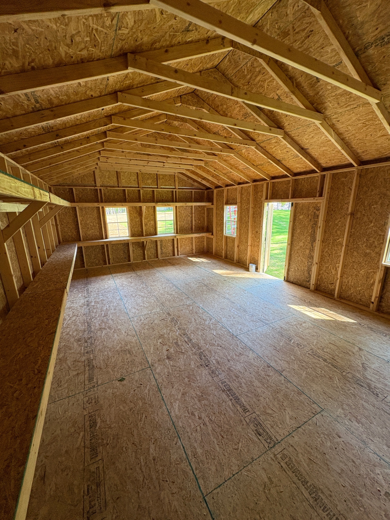 Wide shed interior with open trusses, shelving along the walls, several windows and a house door letting in light