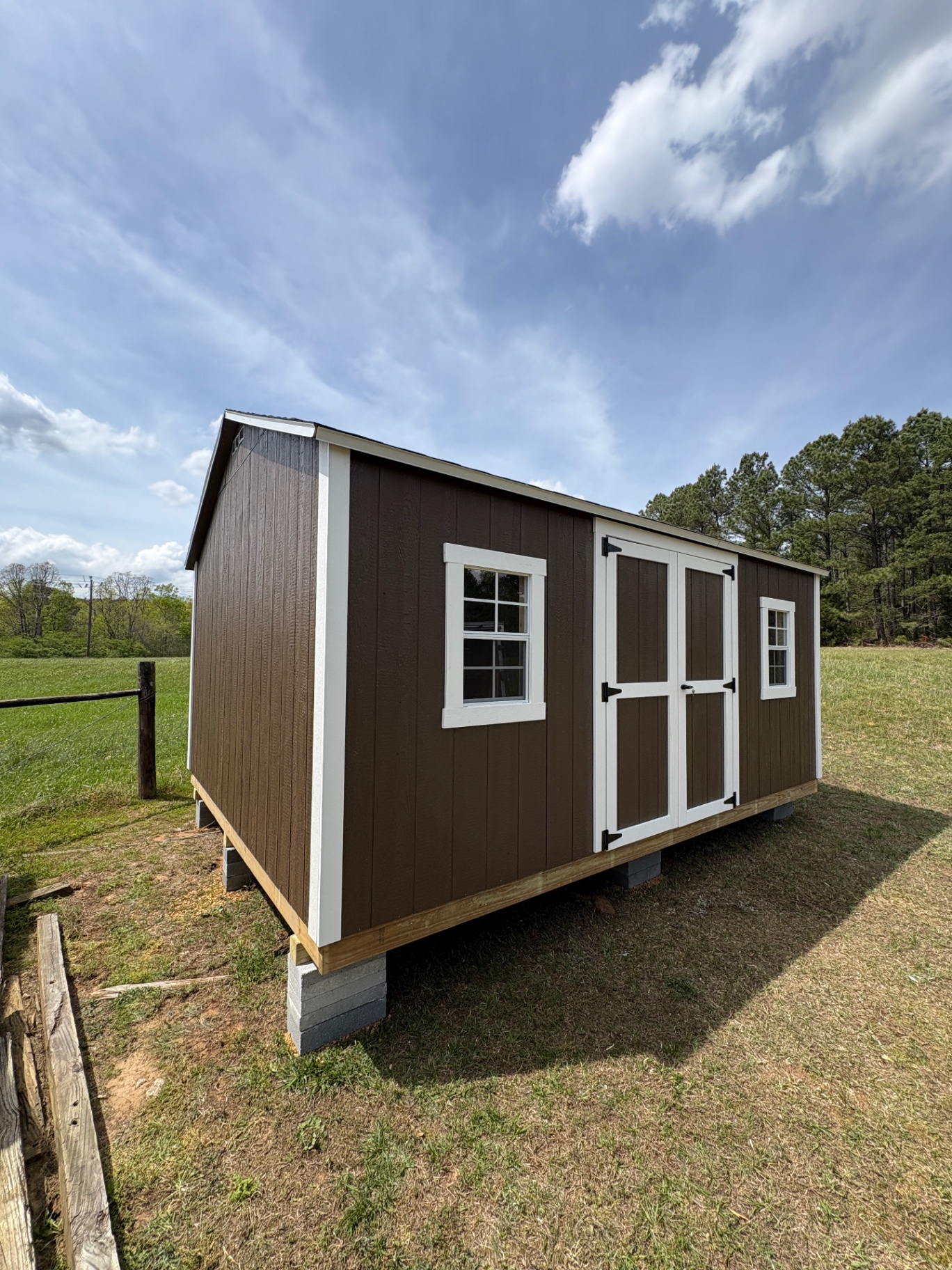 Angled view of a brown shed with white-trimmed double doors and two grid windows in an open field