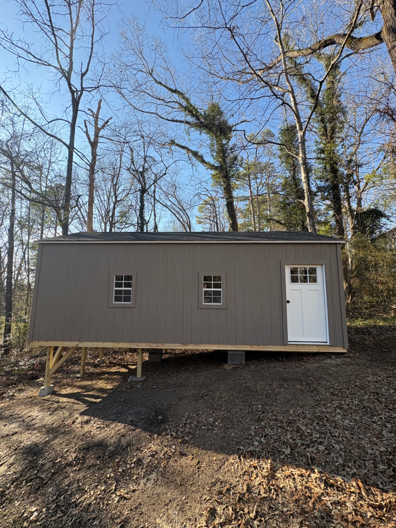 Taupe shed built level on treated posts over a sloped wooded lot, with two grid windows and a white craftsman door