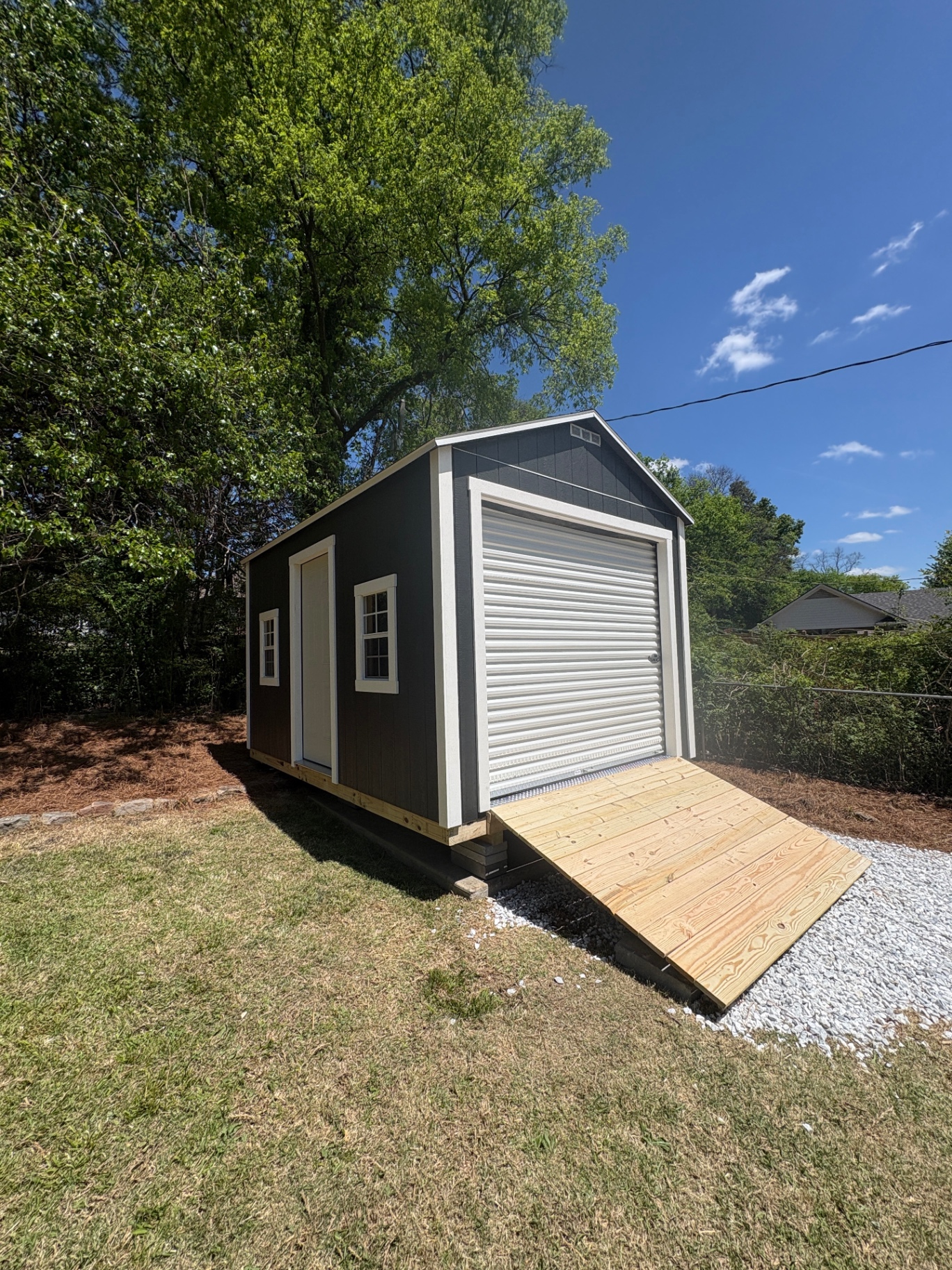 Charcoal shed with a white roll-up garage door, side house door and two windows, with a wide wood ramp over gravel