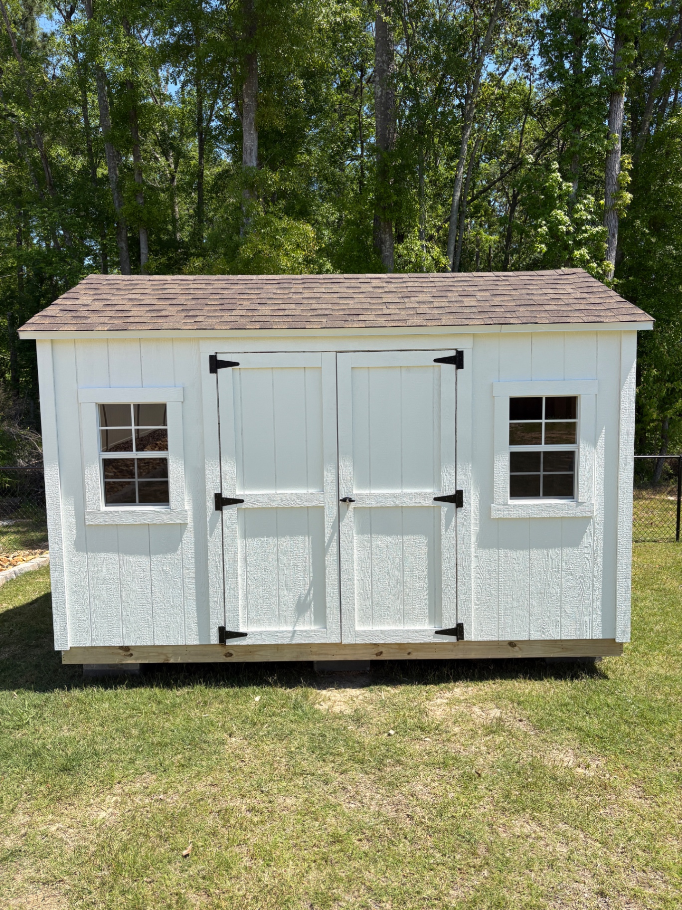White shed with a brown shingle roof, black hinges on double doors and a grid window on each side