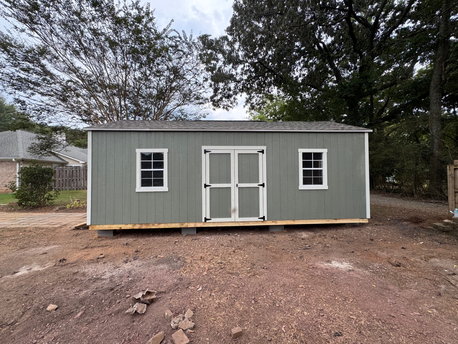 Sage green shed with white trim, centered double doors with black strap hinges, and a grid window on each side