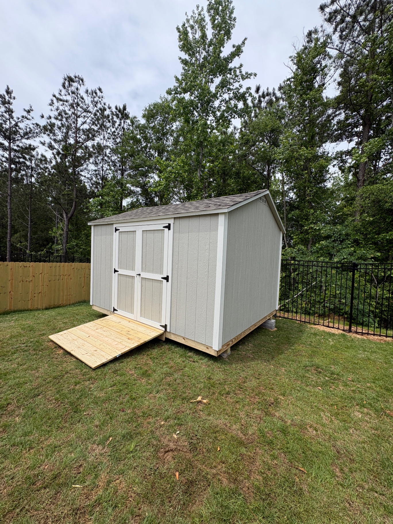 Angled view of a light gray shed with white double doors, gable vent, shingle roof and a wood ramp beside a wood fence