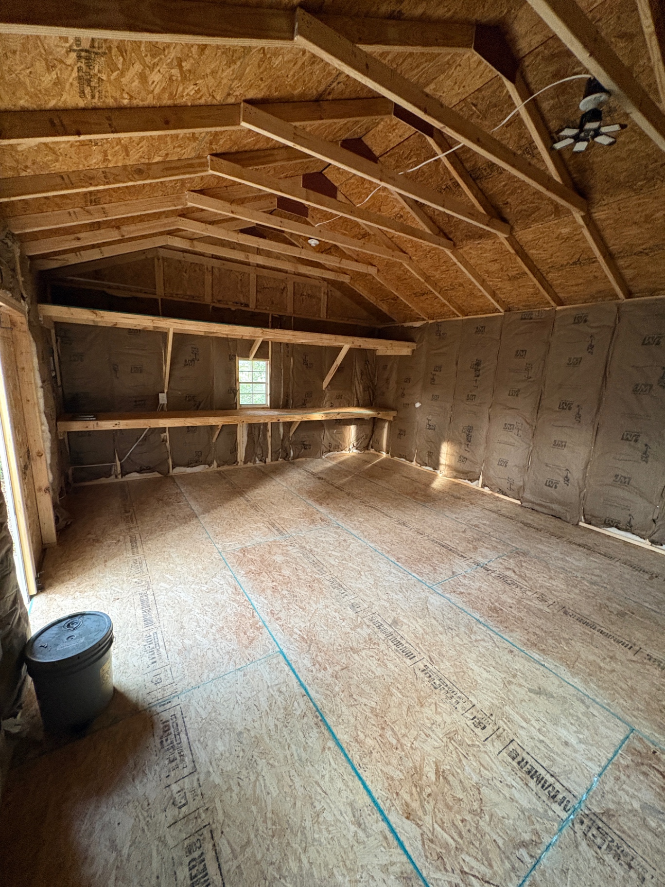 Shed interior with insulated walls, open rafters, a ceiling light, a long shelf and a window