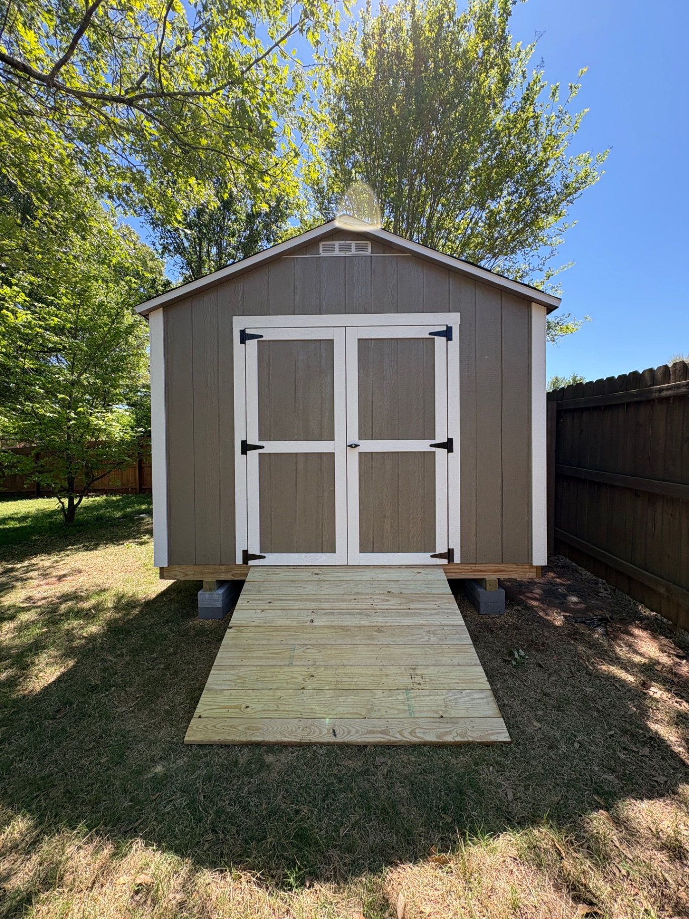 Taupe shed with white-trimmed double doors, a gable vent and a wood ramp, set beside a wood privacy fence