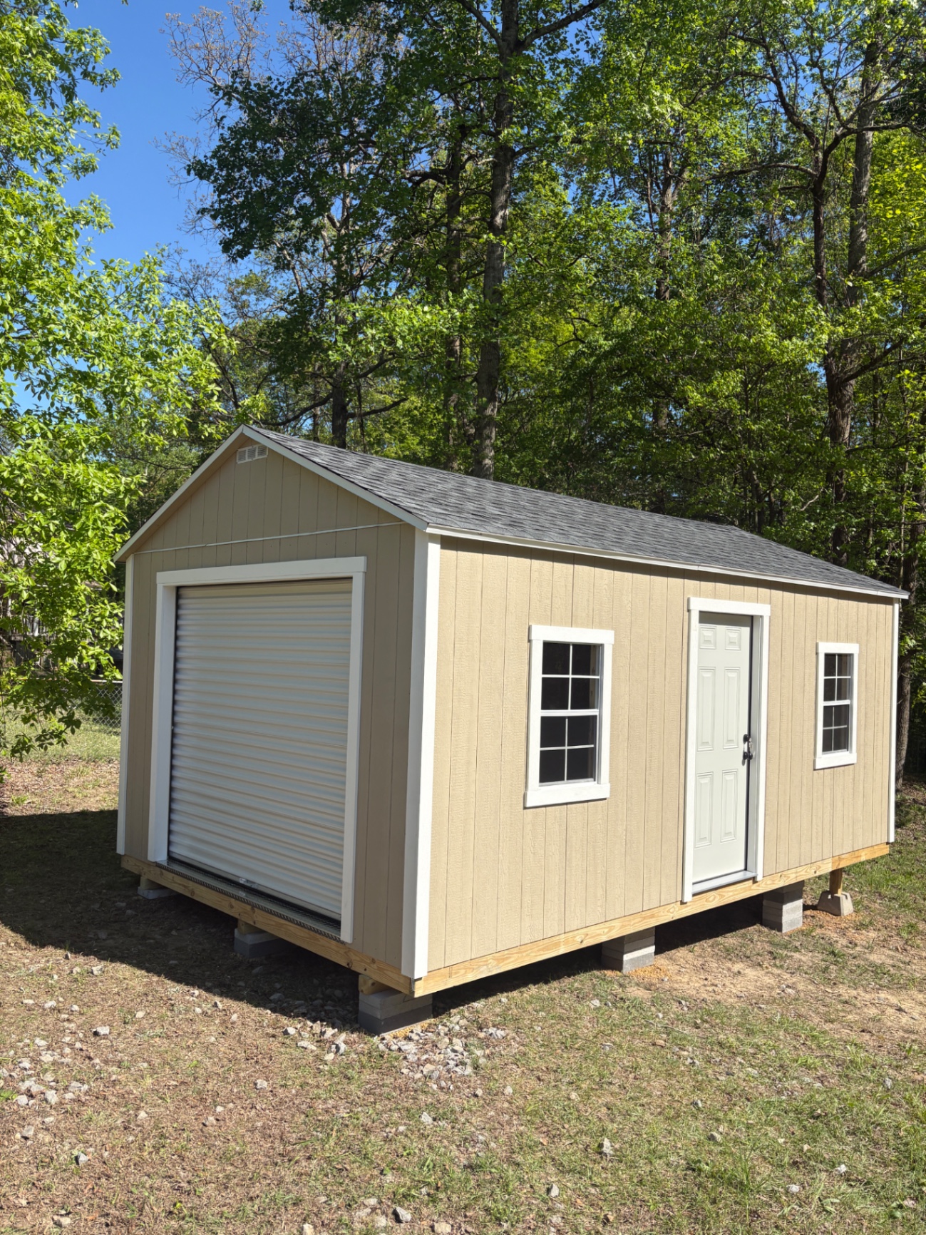 Tan shed with a white roll-up garage door on the gable end, a house door and two grid windows along the side