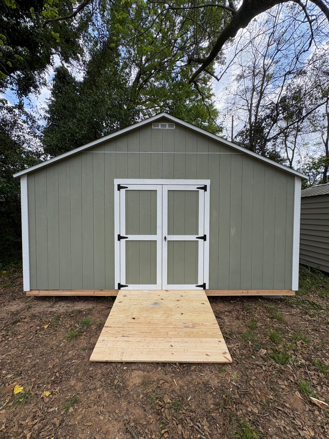 Sage green shed with white double doors, a gable vent and a wide wood ramp at the doors