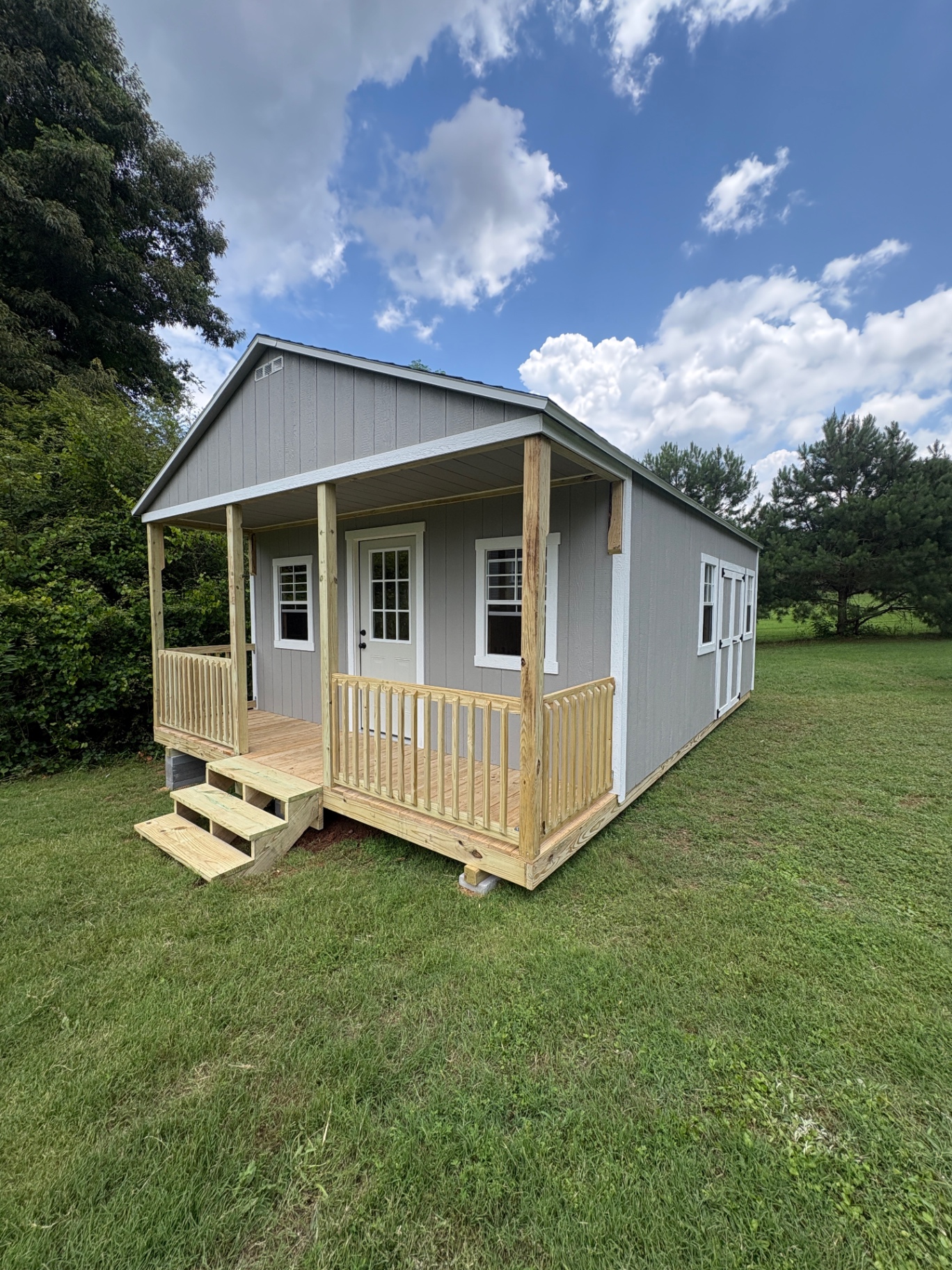 Angled view of a long gray shed with a covered front porch, railing, steps, house door, windows, and double doors on the far end