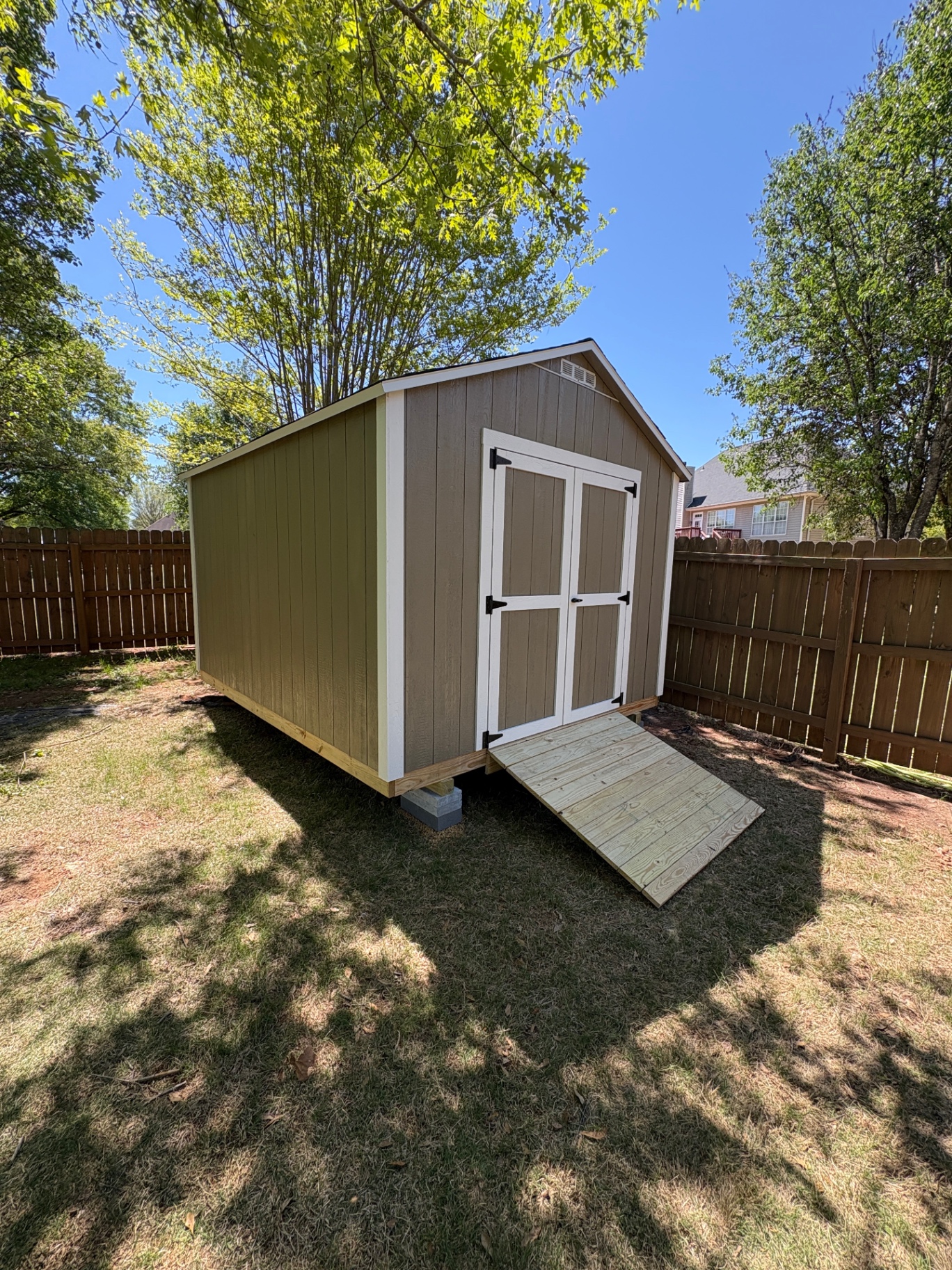 Tan shed with white-trimmed double doors and a wood ramp, set on blocks in a fenced backyard