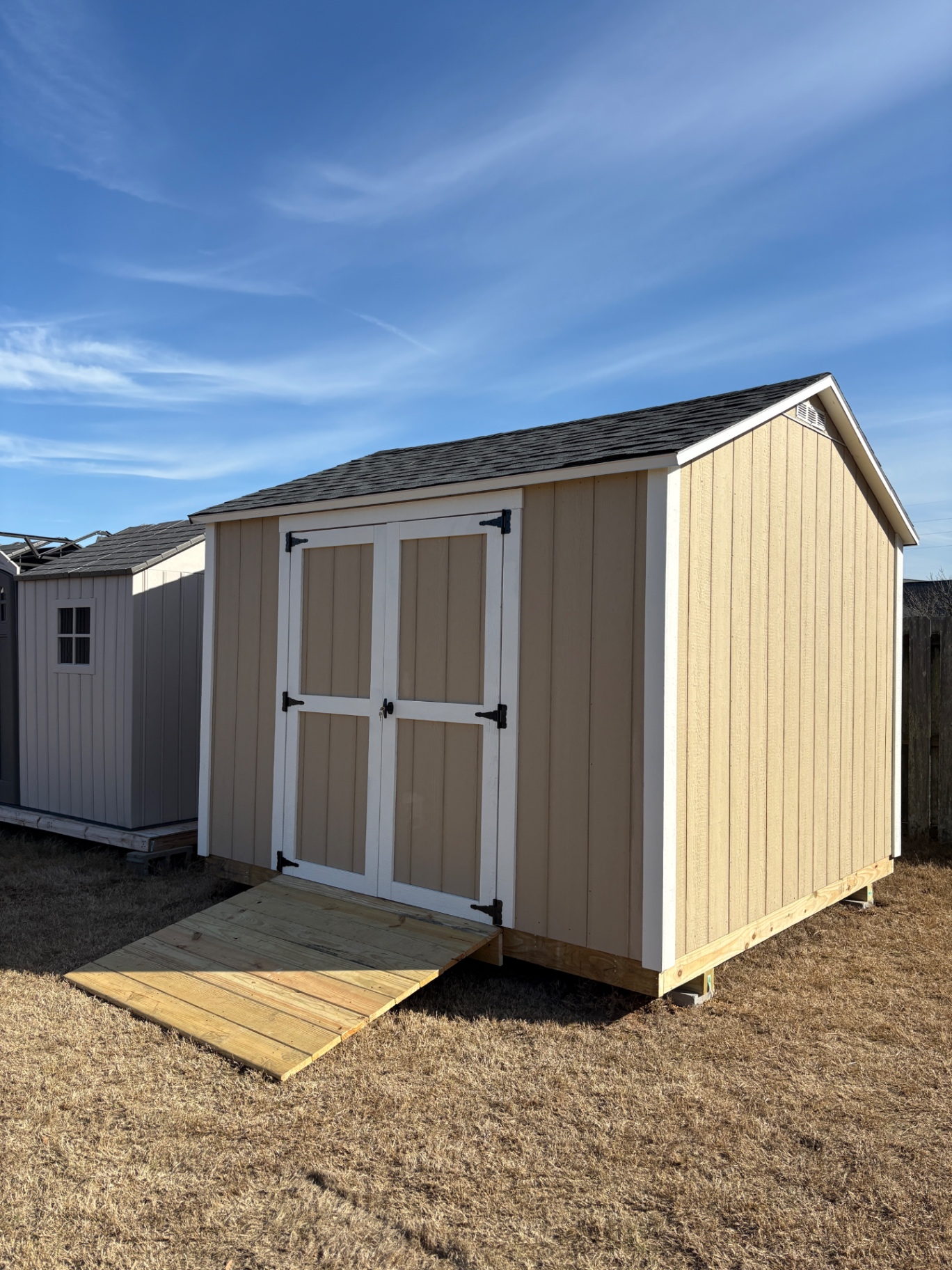 Angled view of a tan shed with white-trimmed double doors, a gable vent and a treated wood ramp