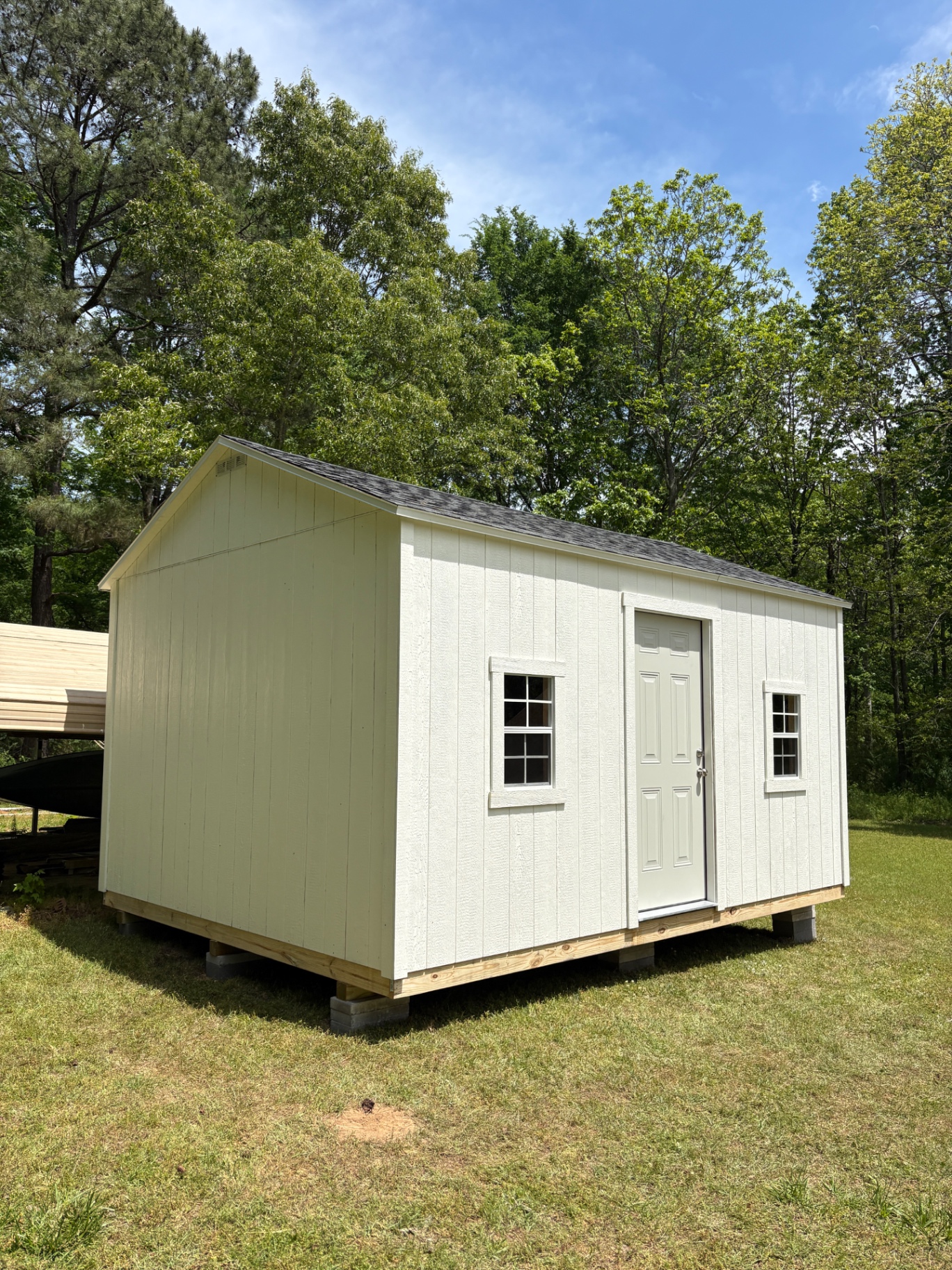 Tan shed with a white house door centered between two grid windows and a shingle roof