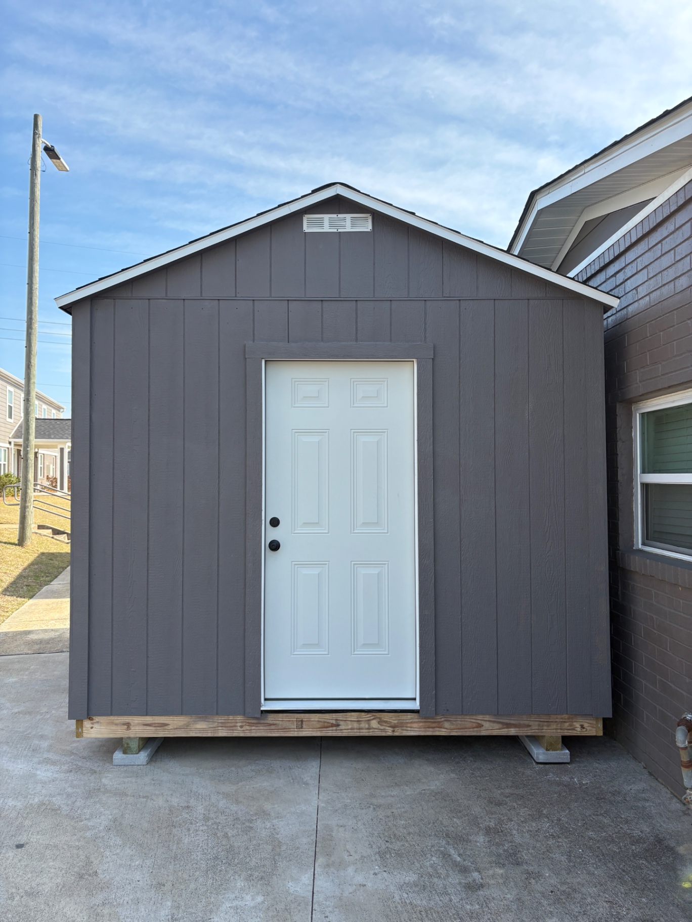 Charcoal shed with a white six-panel house door centered on the gable end and a gable vent above it, set on concrete blocks beside a brick home