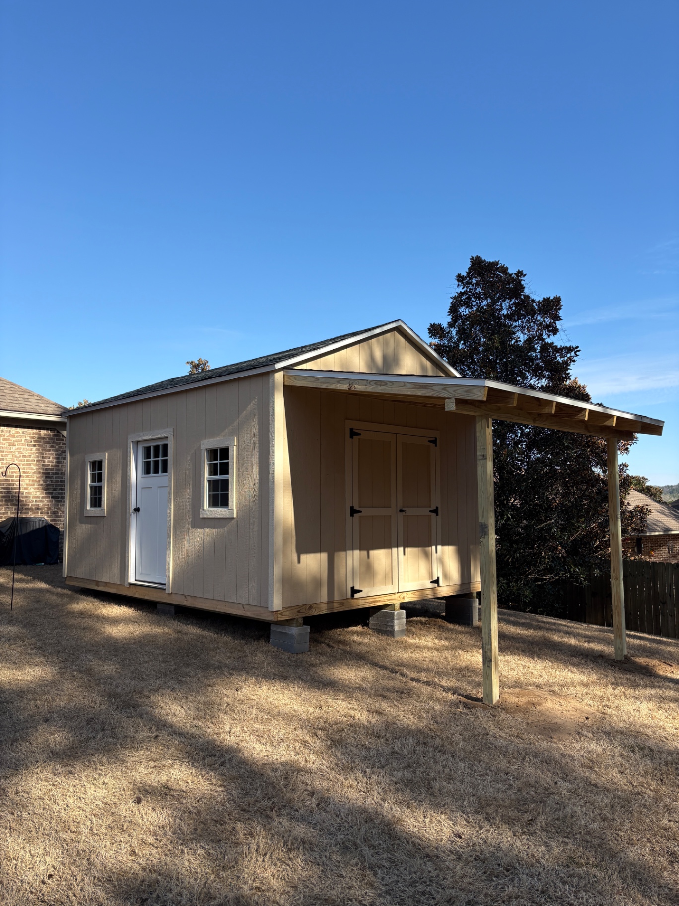 Tan shed with a covered lean-to on posts, double barn doors under the lean-to, a house door with glass and two windows on the long wall