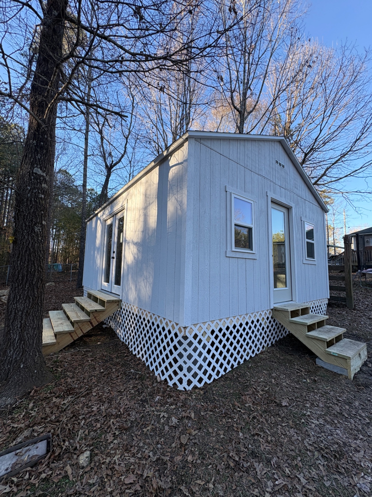 Corner view of a white shed with French doors and a house door on the long wall, standard windows and lattice skirting with steps at both entries