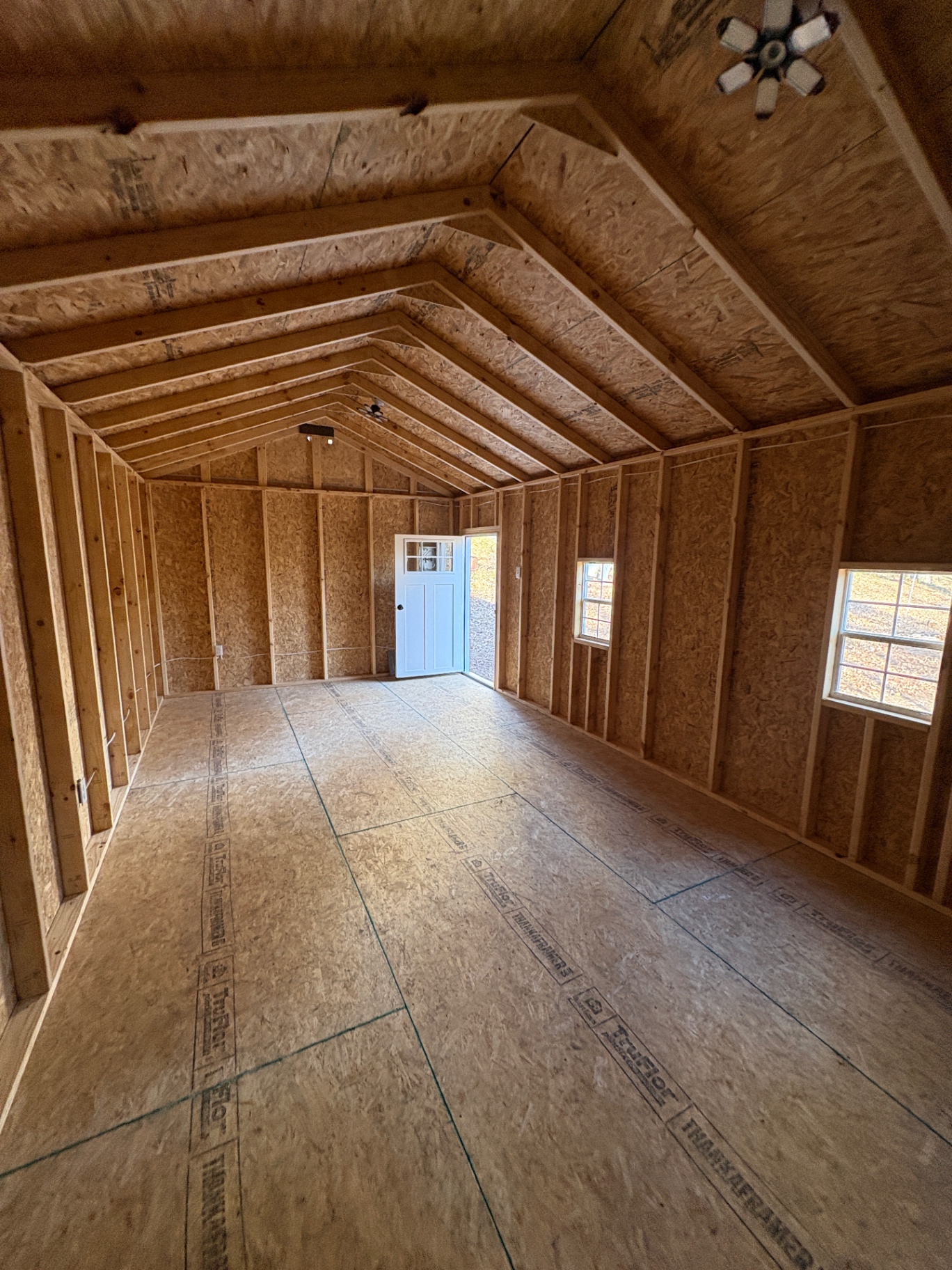 Inside a shed looking toward an open house door, with two ceiling lights, side windows and a TruFloor tongue-and-groove floor