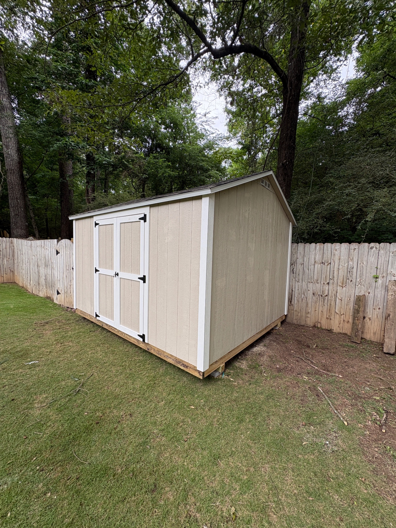 Angled view of a tan shed with white double doors set along a fence line in a shaded backyard