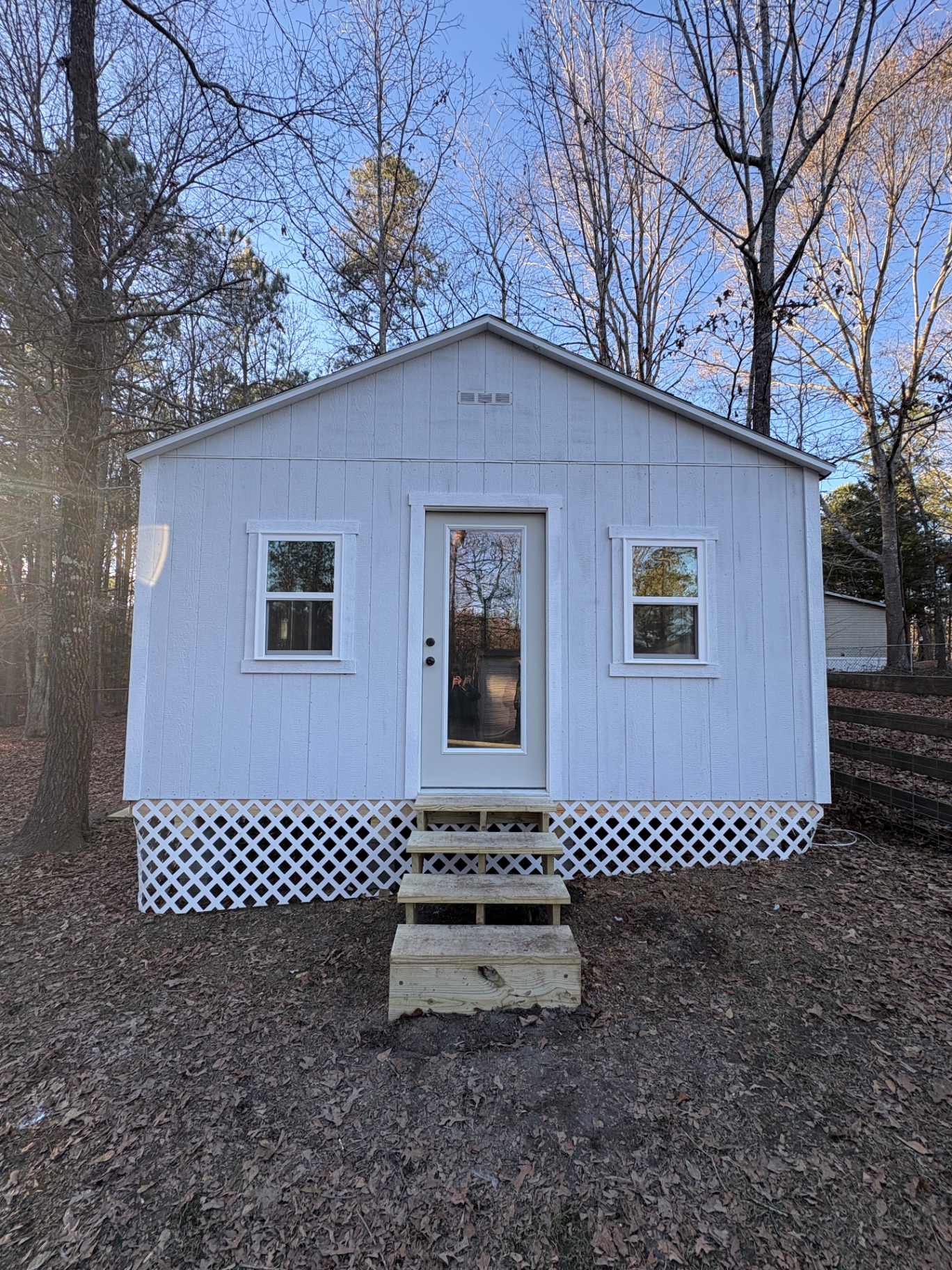 White shed with a full-glass house door, a standard window on each side, a gable vent and white lattice skirting with wood steps