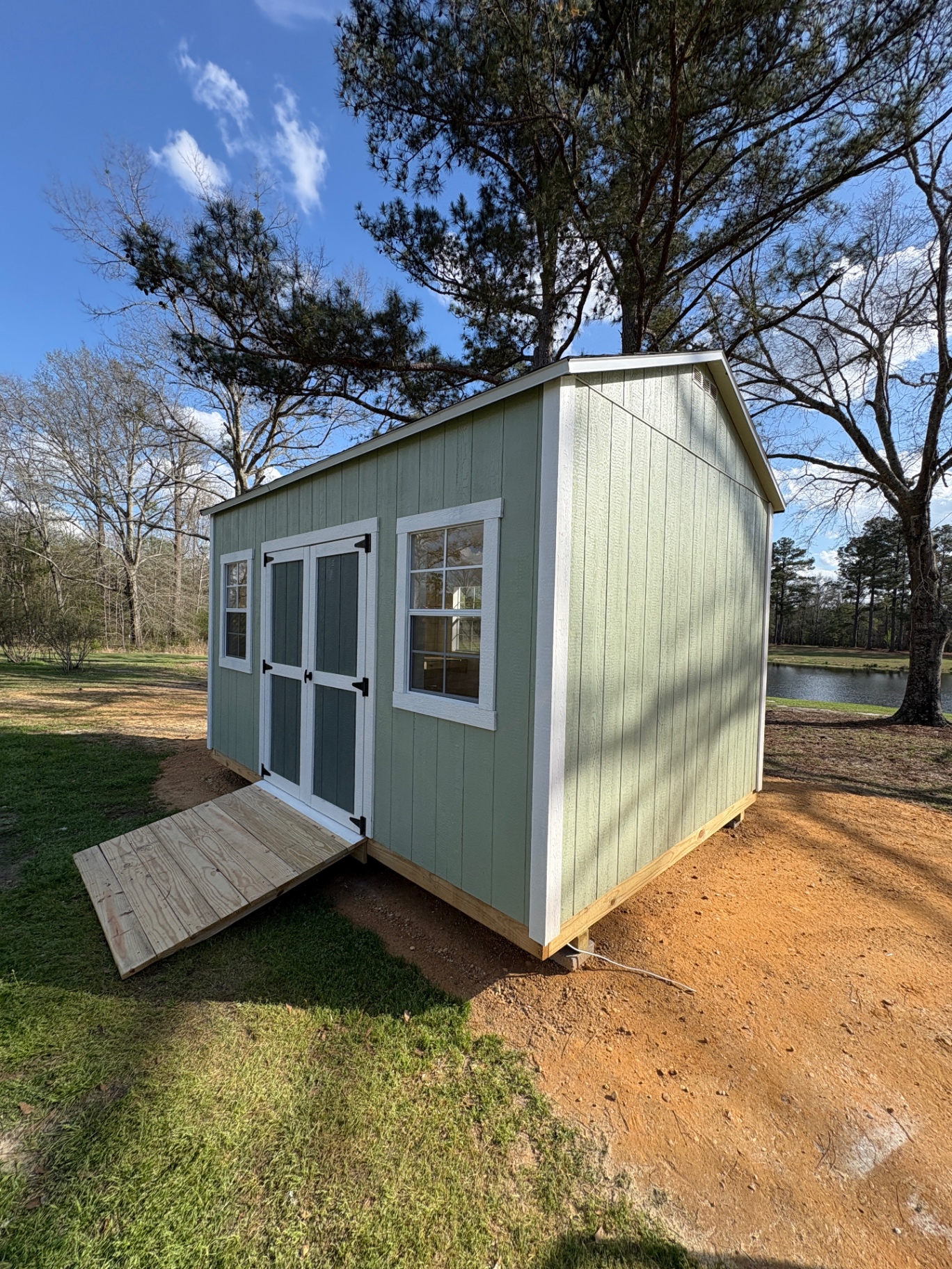 Sage green shed with white-trimmed double doors, a grid window on each side and a treated wood ramp built to fit the doors