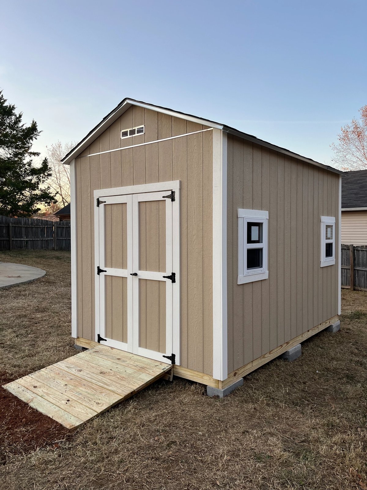 Tan shed with white double doors on the gable end, two side windows, a gable vent and a treated wood ramp