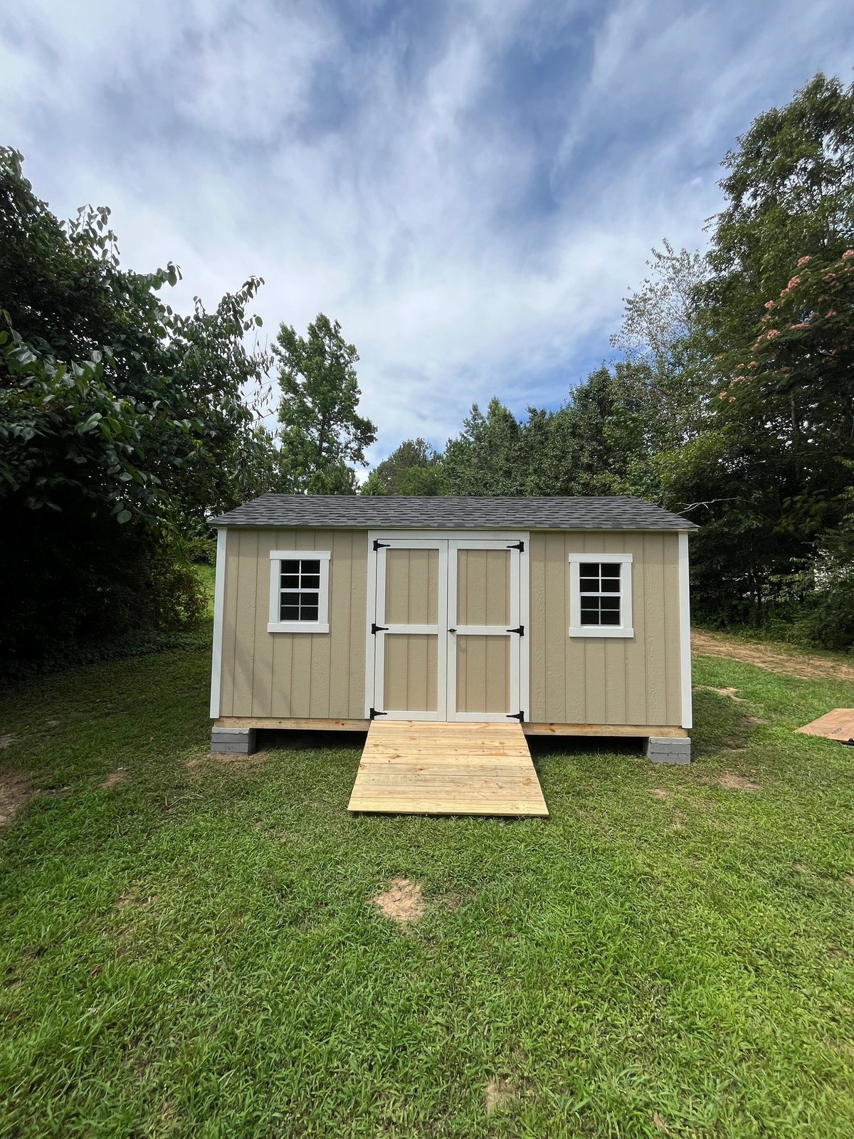 Tan shed with white double doors, two standard windows and a treated wood ramp in a wooded backyard