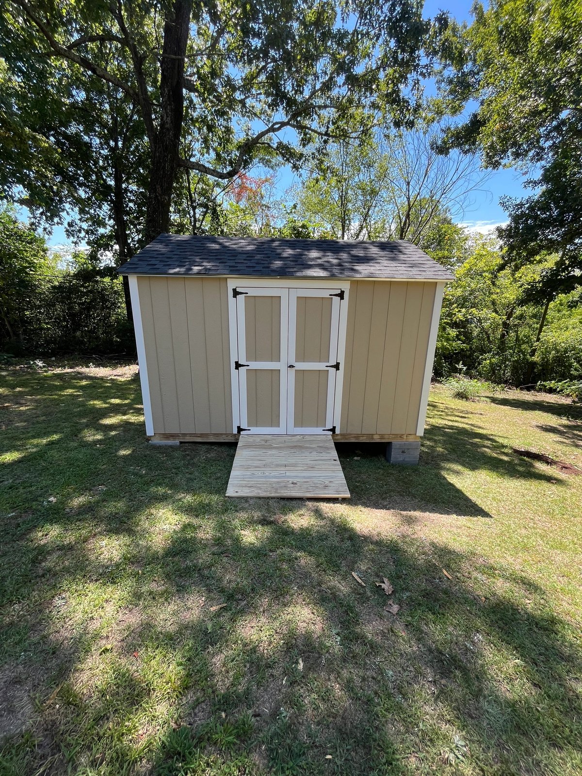 Small wheat-colored shed with white double doors and a treated wood ramp in a shaded yard